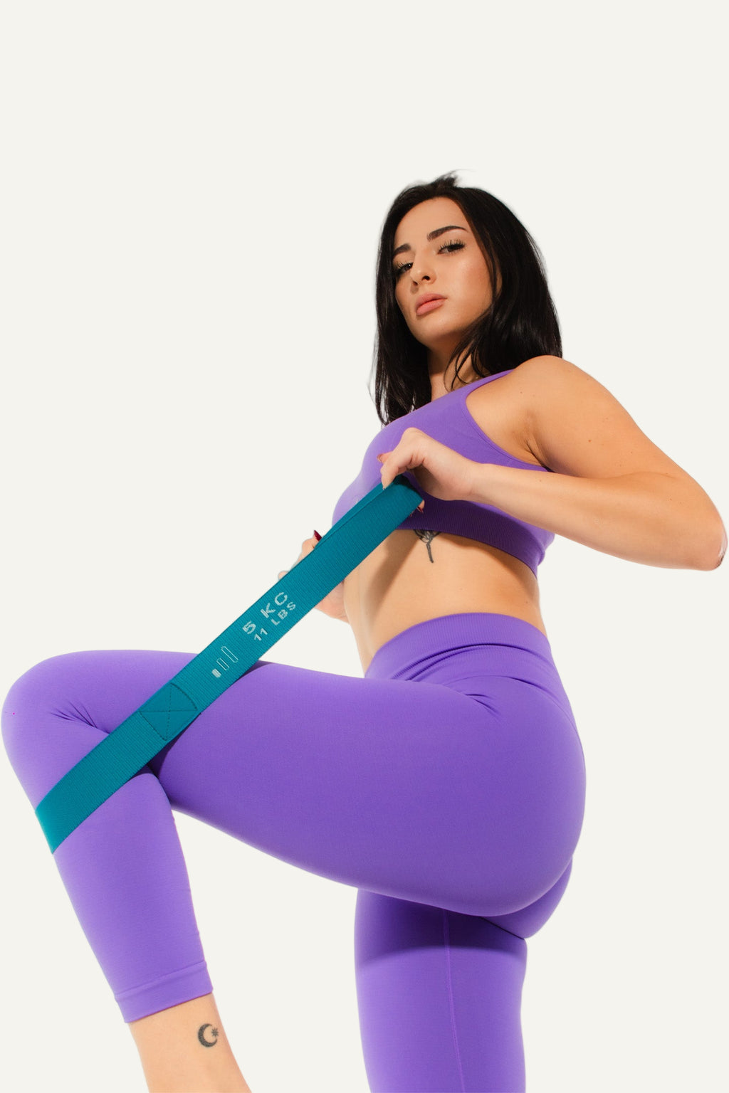 Woman in purple athletic wear using a teal resistance band against a white background